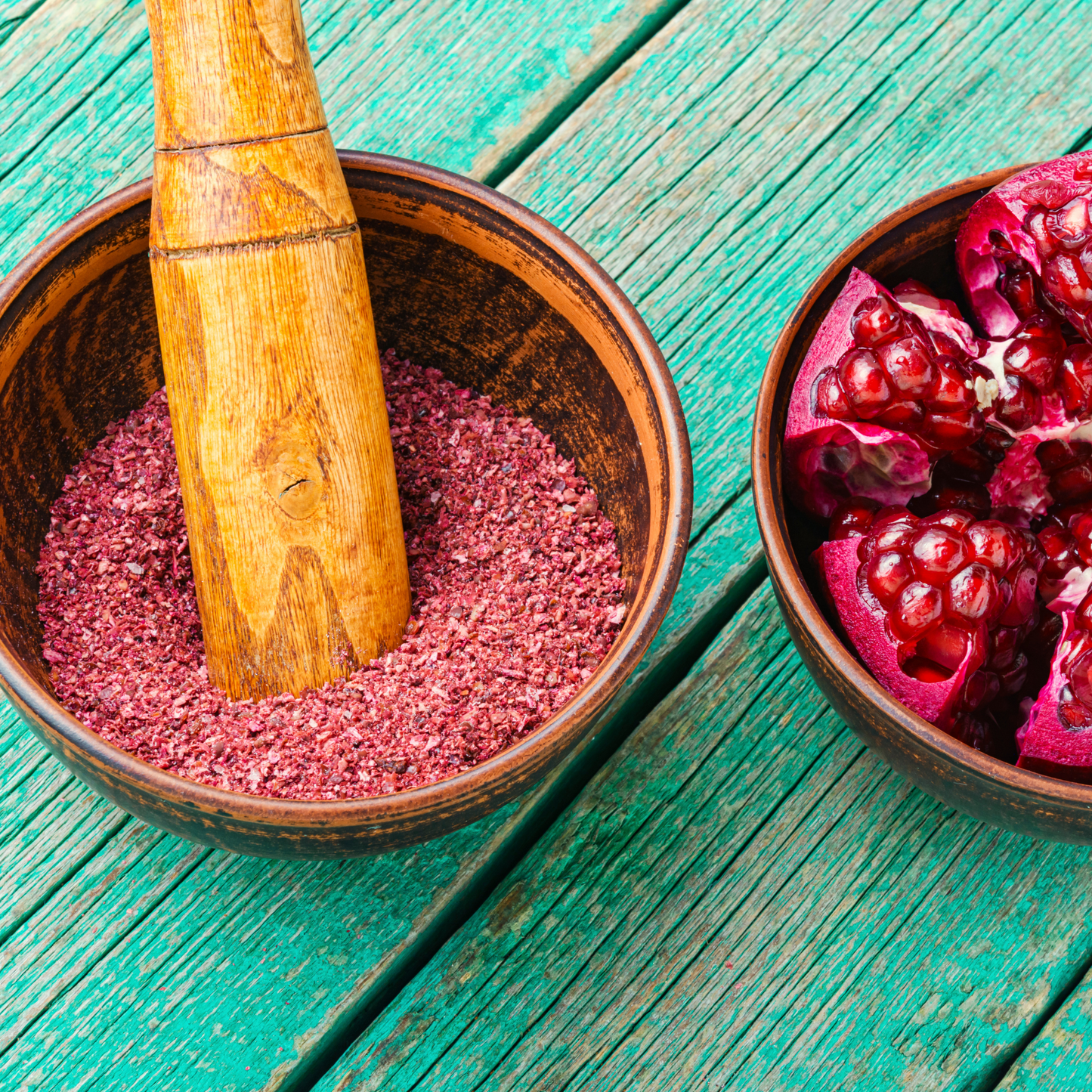 Wooden mortar and pestle with pink powder next to pomegranate pieces on a teal wooden surface